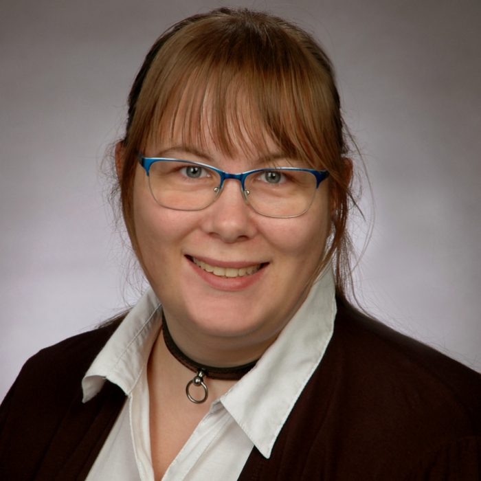 Frau mit blonden Haaren, Brille und Choker, lächelt freundlich in die Kamera.